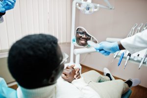 African american man patient in dental chair. Dentist office and doctor practice concept. Professional dentist helping his patient at dentistry medical and showing to him a mirror.